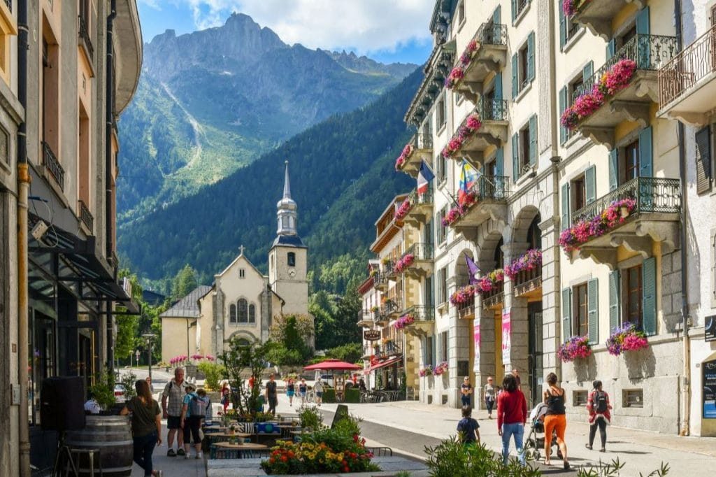Rue animée avec bâtiments fleuris et une église au fond, entourée de montagnes.