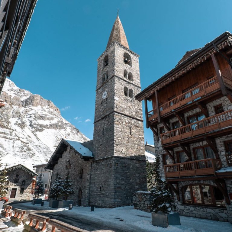 Église en pierre avec un clocher à Val d'Isère, entourée de montagnes enneigées et de chalets.
