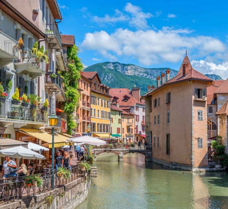 Canal d'Annecy entouré de bâtiments colorés et de montagnes sous un ciel bleu.